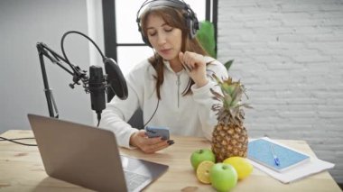 Kulaklık takan genç beyaz bir kadın, elinde akıllı bir telefonla stüdyo mikrofonuna konuşuyor, bir radyo sunucusunu ya da kapalı mekan yayıncılığını canlandırıyor..
