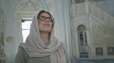 Başörtüsü takan ve gözlüklü genç bir kadın caminin içinde gülümsüyor, kültürler arası memnuniyeti somutlaştırıyor.