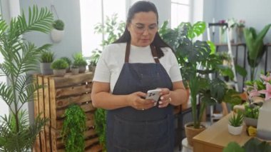 İspanyol kadın çiçekçide akıllı telefon kullanıyor. Etrafı bitki ve buketlerle çevrili.