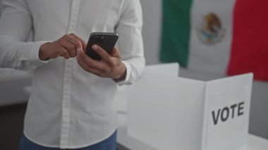 Genç bir İspanyol, arka planda Meksika bayrağı olan bir oy kabininde akıllı telefon kullanıyor..