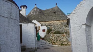 Alberobello, İtalya 'nın büyüleyici sokaklarını keşfeden yeşil elbiseli genç bir kadın, açık mavi gökyüzünün altında ikonik trulli evler ve kaldırım taşından yollar ile çevrili..