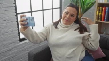 Keyifli orta yaşlı bir kadın sıcak bir oturma odasında akıllı telefonuyla selfie çekerken başparmağını kaldırıyor..
