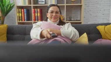 Olgun bir İspanyol kadın, rahat bir oturma odasındaki koltukta uzaktan kumandayla rahatlar..