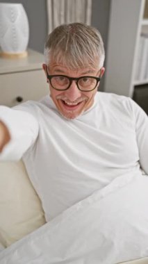 Neşeli gri saçlı orta yaşlı bir adam, pijamasıyla kendinden emin bir poz veriyor, odasında sevinçle gülümsüyor, seni ve kamerayı işaret ediyor.!