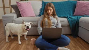 Genç sarışın bir kadın içeride yerde oturuyor, dizüstü bilgisayar kullanıyor ve yakınlardaki beyaz bir köpeğe işaret ediyor..