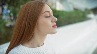 Beyaz kazaklı genç kızıl kadın yeşil şehir parkında gözlerini meditasyon yaparak kapatıyor..