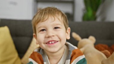 Sarışın çocuk rahat bir oturma odasında oyuncaklarla oynuyor, neşe ve çocukluk masumiyetini ifade ediyor..