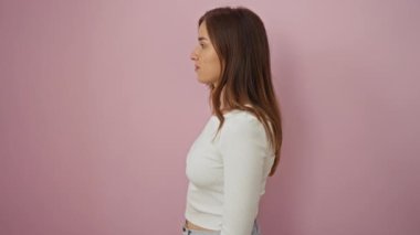 Yan profilde duran kendine güvenen genç sarışın kadın doğal bir yüz, rahat bir gülümseme, soyutlanmış pembe arka planda mutlu ve neşeli davranışlar sergiliyor.