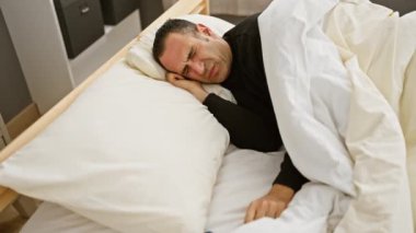 Beyaz çarşaflı, sıkıntılı, siyah gömlekli İspanyol bir adam modern bir yatak odasında rahatsız bir şekilde dinleniyor..