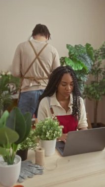 Bir kadın ve bir erkek kapalı bir çiçekçide birlikte çalışıyor. Dişi, dizüstü bilgisayar kullanıyor ve erkek bitkileri düzenliyor..