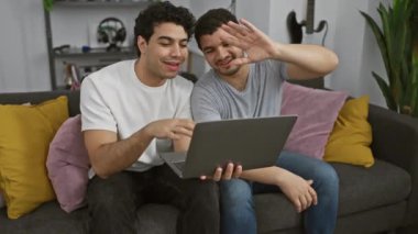 İki adam gülümsüyor ve arka planda renkli yastıklar ve gitarla rahat bir oturma odasında dizüstü bilgisayar kullanıyor.