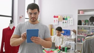 İki adam terzi dükkanında çalışıyor, biri dikiş dikerken diğeri de mankenlerin ve kumaşların arasında tablet kullanıyor..