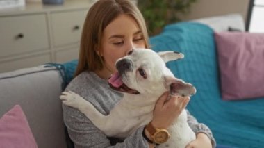 Genç bir kadın, rahat bir oturma odasında beyaz köpeğini öpüyor ve evcil hayvan ve sahibi arasında hassas bir an olduğunu gösteriyor..