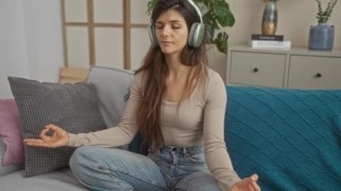 Evdeki kanepede meditasyon yapan, kulaklık takan ve rahat bir oturma odasında günlük giysiler giyen esmer, çekici İspanyol bir kadın..
