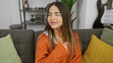 Neşeli genç Latin kadın, neşeyle parıldıyor, rahat oturma odasında oturuyor, baş parmakları havada ve rahat süveteri içinde, ışıl ışıl gülümseyen bir şekilde kenarı işaret ediyor..