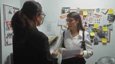 İki kadın soruşturma posterleri ve belgelerle dolu bir odada bir davayı tartışıyor ve bir dedektiflik bürosu ortamı yaratıyor..