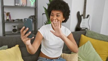 Neşeli bir Afro-Amerikalı kadın evinde video görüşmesi yapıyor, modern bir oturma odası arka planı gösteriyor..