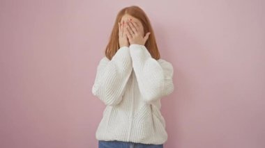 Depresif bir kızıl, genç bir kadın, süveter giymiş, gözyaşları içinde yüzünü kapatıyor, üzgün ifadesi pembe bir arka planda izole edilmiş.