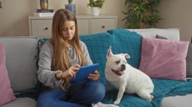 Genç bir kadın, rahat bir oturma odasında, kanepede köpeğiyle otururken tabletle etkileşime giriyor..