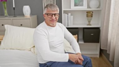 Pijamalı neşeli gri saçlı adam, yatak odasında sıcak bir kucaklama için kollarını açıyor. Saf mutluluğun geniş, misafirperver gülümsemesi kameraya bakarken yüzünü aydınlatıyor..