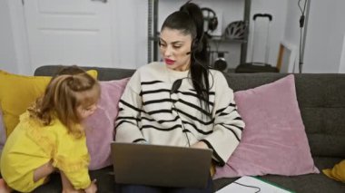 Kulaklıklı bir kadın evde dizüstü bilgisayar kullanırken sarı elbiseli genç kızı yanında oturuyor..