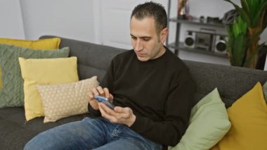 Neşeli İspanyol bir adam akıllı telefonunu renkli yastıklarla çevrili bir kanepede, rahat bir oturma odasında kullanır..
