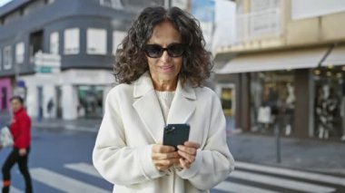 Şehirde akıllı telefon kullanan güneş gözlüklü ve ceketli olgun bir İspanyol kadın.