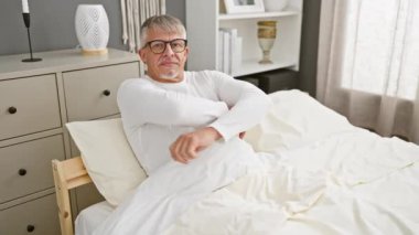 Neşeli orta yaşlı adam, kollarını kavuşturmuş, yatak odasında kendine güvenle parıldıyor, gri saçları gülümsemesini övüyor.