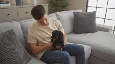 Oturma odasında oturan genç bir adam elinde köpeğiyle gri bir kanepede oturuyor..