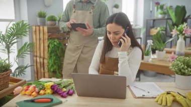 Bir kadın telefon görüşmesi ve dizüstü bilgisayarda dijital tabletli bir adam olarak yoğun bir çiçekçide asistanlık yapıyor..
