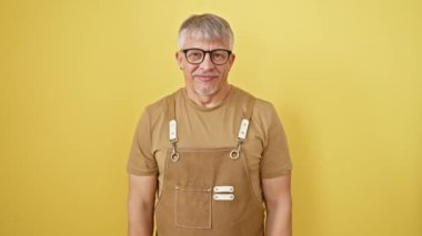 Gözlüklü ve önlüklü neşeli gri saçlı orta yaşlı adam, sarı izole bir arka planda sana neşeli bir gülümseme ve sevinçle bakıyor. Mutlu titreşimlere hizmet etmek, dik durmak, iyi enerji yaymak..