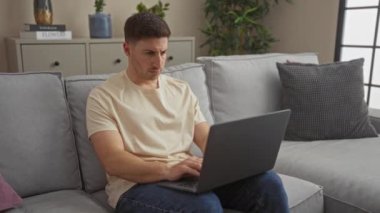 Genç bir İspanyol, oturma odasındaki gri bir kanepede otururken dizüstü bilgisayar kullanıyor. Rahat bir ev ortamında derinden konsantre oluyor..