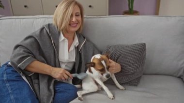 Orta yaşlı sarışın bir kadın oturma odasında küçük köpeğini tararken gri bir kanepede oturmuş modern dekorlu rahat bir ev atmosferi sergiliyor..