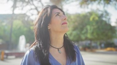 Olgun İspanyol kadın güneşli bir park ortamında doğayı düşünür. Huzur ve yansıma hissi verir..