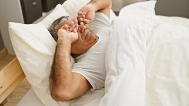 Beyaz tişörtlü gri saçlı bir adam rahat bir yatak odasında uykusundan uyanırken esniyor ve sabah rutinini anımsatıyor..