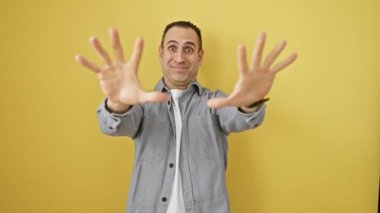 Kendinden emin ve mutlu İspanyol bir adam, tişörtlü, gülümseyen ve gururla '10' luk elini gösteren güneşli sarı bir zeminde sayma ve haberleşme işareti yapıyor.