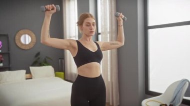 Orta karar dekore edilmiş bir yatak odasında dambıllarla egzersiz yapan genç bir kadın evde spor yapıyor..