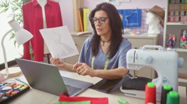 Olgun bir İspanyol kadın moda tasarımcısı, terzi dükkanında bir video görüşmesi yapıyor, dikiş ekipmanlarıyla bir elbise tasarımı sergiliyor..