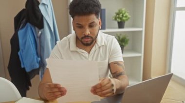 Kafası karışmış bir Afrikalı Amerikalı, arka planda dizüstü bilgisayarı ve dekoru olan bir ofiste evrak okuyor..