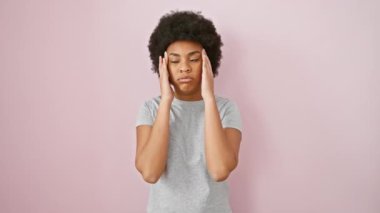 Stresli Afrikalı Amerikalı kadın, el ele tutuşmuş, baş ağrısı ve migren ağrısı çekiyor. Tişört giyiyor, pembe arka planda tek başına duruyor.