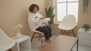 Kıvırcık saçlı olgun bir kadın oturma odasında beyaz bir chihuahua köpeği tutuyor..