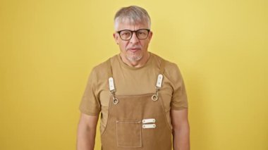 Tiksindirici orta yaşlı, gri saçlı, önlüklü, gözlüklü, izole edilmiş sarı arka planda duran, dayanılmaz, kokuşmuş bir kokuya tepki olarak burnunu çimdikleyen bir adam..