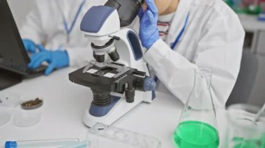 Laboratuvar önlüğü giymiş bir kadın ve bir erkek, bilimsel bir araştırma laboratuvarında mikroskop ve bilgisayarla birlikte çalışıyorlar..