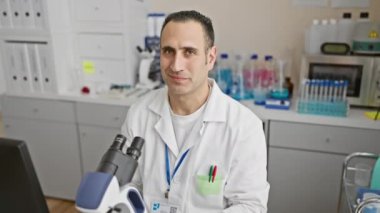 Bilim adamı üniforması içindeki neşeli, genç İspanyol adam, laboratuvarda özenle çalışıyor, güvenle işaret ediyor ve gülümsüyor, pozitif enerji yayıyor..