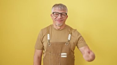 Neşeli orta yaşlı, gri saçlı, gözlüklü ve önlüklü adam, kendinden geçmiş bir şekilde yüzüne ve burnuna bakıyor, parlak sarı izole bir arka planda güzellik sağlığını destekliyor.