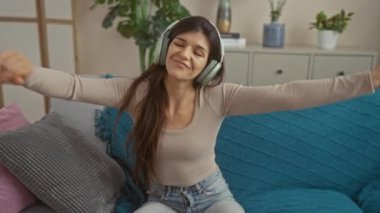 Evdeki rahat bir oturma odasında kanepede otururken, kulaklıkla müzik eşliğinde mutlu bir şekilde dans eden genç İspanyol kadın..
