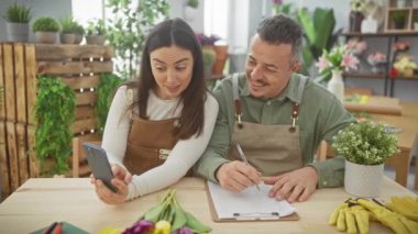 Çiçekçi dükkanında önlüklü bir adam ve bir kadın çiçek buketleriyle dolu bir pano üzerinde etkileşim halindeler..
