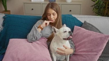 Evde uzun sarı saçlı genç bir kadın, oturma odasında bir kanepede oturuyor, pembe ve mavi yastıklarla çevrili beyaz köpek evcil köpeğine sarılıyor..