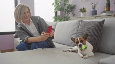 Orta yaşlı bir kadın oturma odasında oturmuş köpeğinin kırmızı akıllı telefonuyla fotoğraflarını çekiyor. Köpek de tenis topuyla oynuyor..