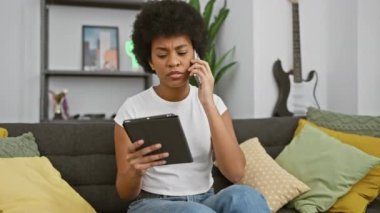 Afro-Amerikan kadın evde tablet ve telefon ile birden fazla iş yapıyor, ev bitkileri ve modern oturma odası dekoru ile çevrili..